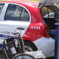 A Yad Sarah hatchback vehicle in bright red, white and blue, hatchback open with driver adjusting specialized bed and wheelchair