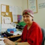 Dr. Shlomit Lehman of the Family Centre Negev sits behind her desk. She is wearing a red sweater, has her head covered with a small hat and is smiling at the camera