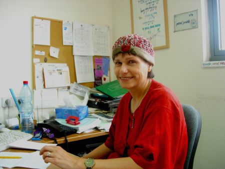 Dr. Shlomit Lehman of the Family Centre Negev sits behind her desk. She is wearing a red sweater, has her head covered with a small hat and is smiling at the camera